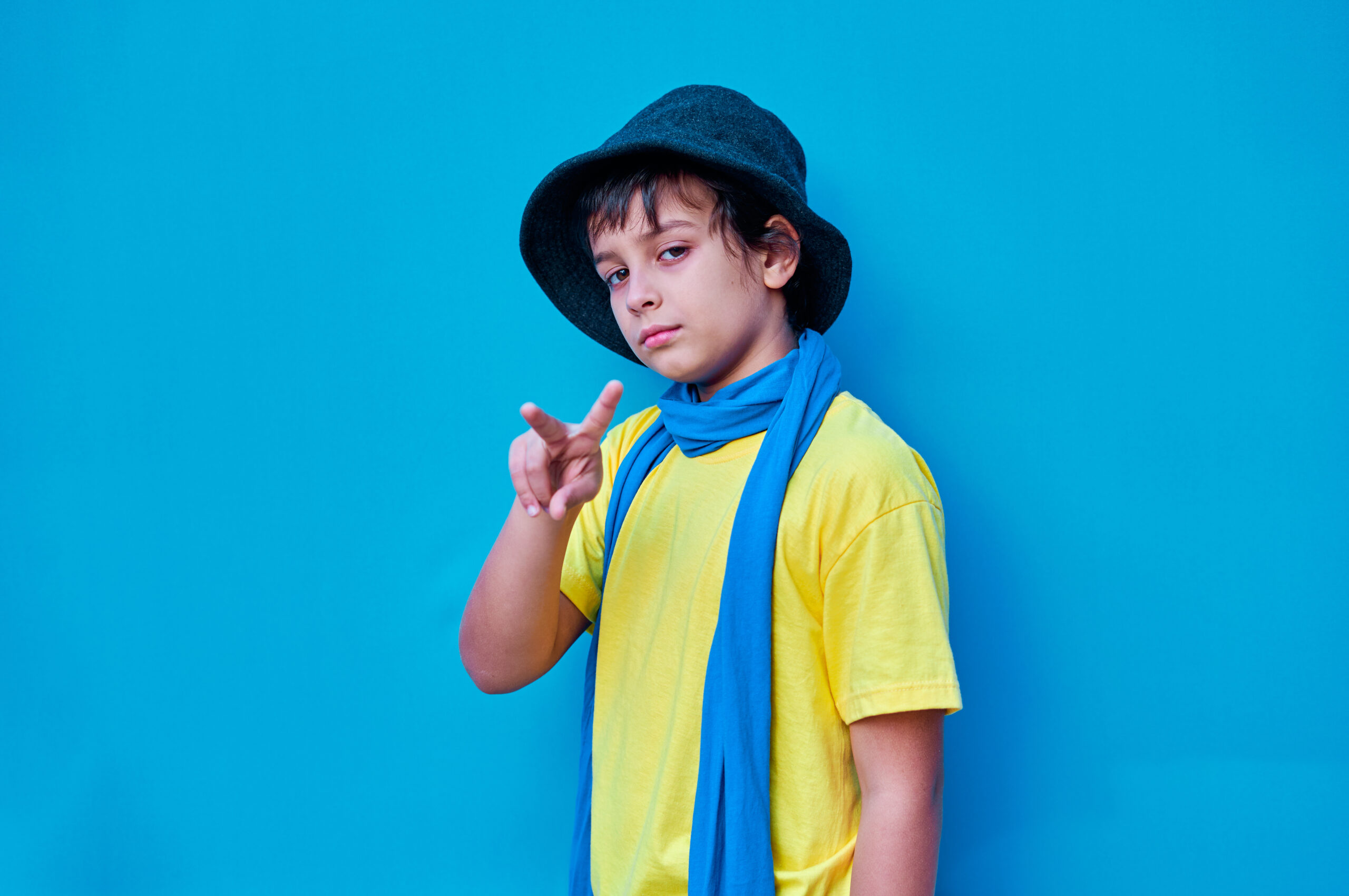 A Portrait of defiant boy in yellow t-shirt, scarf and hat doing, on blue background. Copy space